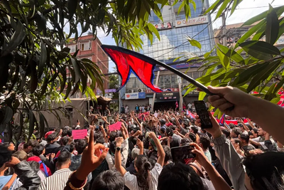 A large, energetic crowd of young demonstrators gathers on a city street to voice their concerns. Participants wave flags and raise their hands in unity during a passionate public rally.