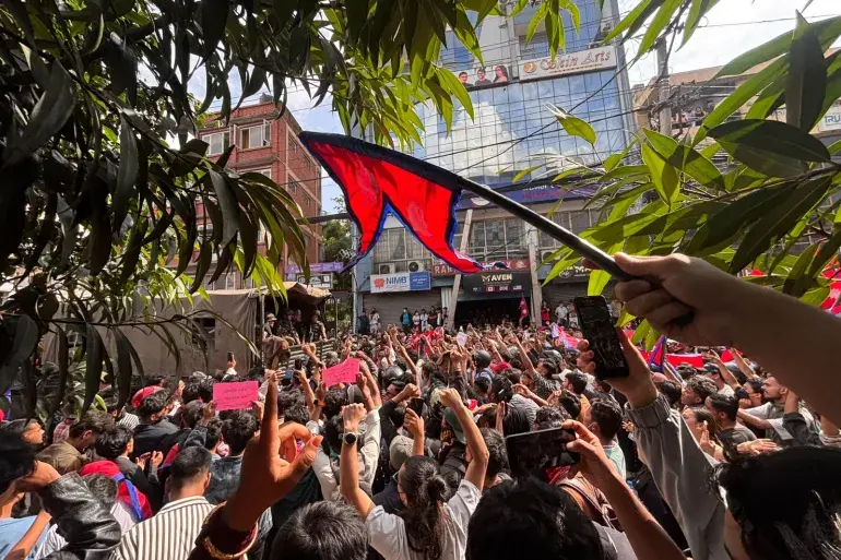 A large, energetic crowd of young demonstrators gathers on a city street to voice their concerns. Participants wave flags and raise their hands in unity during a passionate public rally.